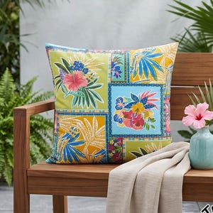 May include: A square throw pillow featuring a patchwork design of tropical floral patterns in shades of lime green, mustard yellow, and light blue. The print includes hibiscus flowers and palm leaves. The pillow is placed on a wooden bench.