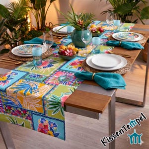 Peut inclure: Un chemin de table rectangulaire orné d'un motif floral tropical aux tons vifs de jaune, vert, bleu et rose. Il est disposé sur une table en bois avec des assiettes blanches, des serviettes en tissu bleu sarcelle, des verres et une coupe de fruits.