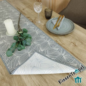 Könnte beinhalten: Ein grauer und weißer Tischläufer mit Muster und grauer Umrandung. Der Läufer ist in der Mitte gefaltet, sodass die Rückseite sichtbar ist, die weiß mit einem grauen Muster ist. Der Läufer liegt auf einem Holztisch mit einem Glas Wasser, einer Kerze und einem Teller mit einer Serviette und Besteck.
