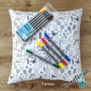 May include: A white square throw pillow cover featuring black line art of unicorns, rainbows, clouds, stars, and flowers. Included is a clear plastic pack of six textile markers in assorted colors, with four additional loose markers.