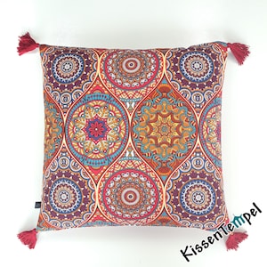 Könnte beinhalten: Quadratisches Kissen mit einem farbenfrohen Mandala-Muster in Rot-, Orange-, Blau- und Lilatönen. Es hat vier rote Quasten an den Ecken. Der Text "KissenTempel" ist unten rechts aufgedruckt.