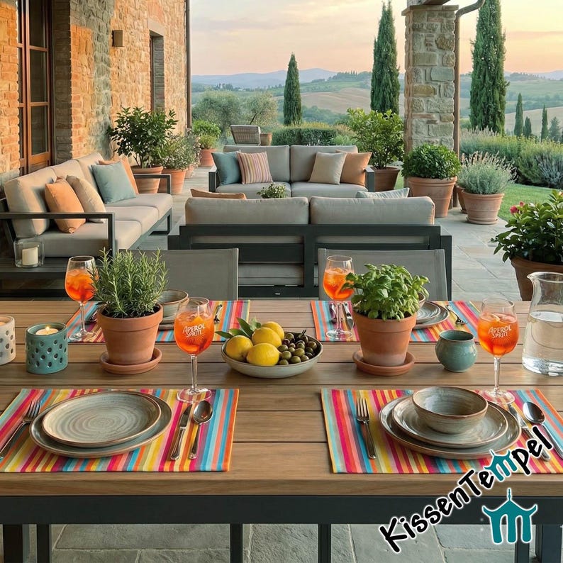 Puede incluir: Escena de comedor al aire libre con una mesa de madera preparada para una comida. Manteles individuales a rayas de colores, platos, vasos con bebidas naranjas y hierbas y plantas en macetas. El fondo muestra un patio con asientos y una vista panor&aacute;mica.