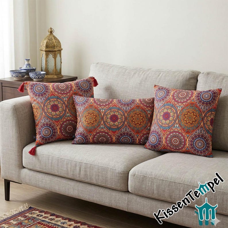 May include: Three decorative throw pillows with a vibrant, circular mandala pattern in shades of red, blue, and gold rest on a gray sofa. Two square pillows and one rectangular pillow are visible, with red tassels on the square pillows.