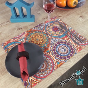 Può includere: Un tovaglietta colorata con un motivo a mandala in tonalità di rosso, arancione, giallo, blu e viola. La tovaglietta è posizionata su un tavolo di legno con un piatto nero, un tovagliolo rosso e un bicchiere di vino.