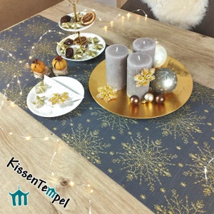 Può includere: Un runner da tavolo grigio con un motivo a fiocco di neve dorato. Il runner è decorato con candele, ornamenti e un vassoio a più livelli con frutta secca e decorazioni a forma di stella. Il testo "KissenTempel" è visibile nell'angolo in basso a sinistra dell'immagine.
