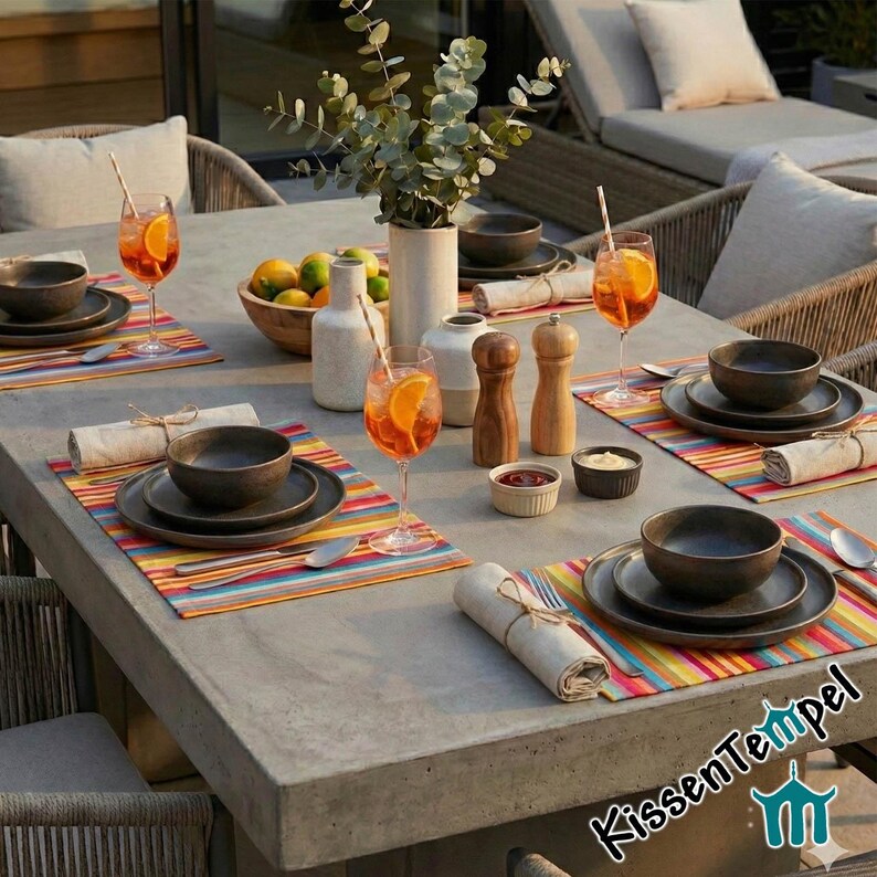 Puede incluir: Mesa para comer al aire libre con cuencos y platos de color gris oscuro sobre manteles individuales a rayas de colores. Incluye c&oacute;cteles de naranja, saleros y pimenteros de madera y peque&ntilde;os cuencos con condimentos. Un jarr&oacute;n con vegetaci&oacute;n y limones completa la escena.