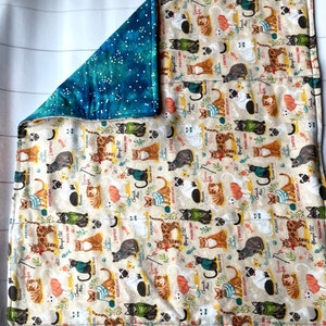 May include: Reversible cat-themed blanket. One side features a beige background with various cat breeds, names, and playful illustrations. The reverse side has a blue background with white star-like patterns. The blanket is rectangular.