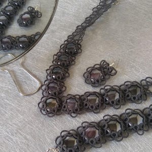 May include: A dark gray jewelry set featuring a necklace, bracelet, and earrings. Each piece has a floral lace design with dark purple-gray gemstones. The set is displayed on a light gray surface, with a mirror reflecting the earrings.