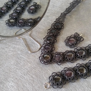 Puede incluir: Un conjunto de collar, pulsera y pendientes de ganchillo negro con detalles de piedras preciosas oscuras. Las joyas se muestran sobre un fondo plateado.