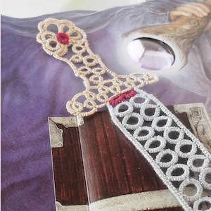 May include: A silver and gold lace bookmark shaped like a sword. The sword has a red jewel on the pommel and a red stripe on the blade.