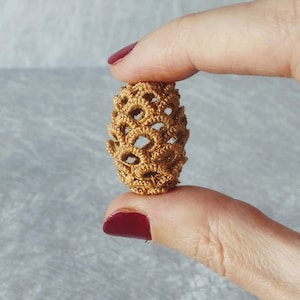 Może przedstawiać: Mały, złotobrązowy, szydełkowy ornament w kształcie szyszki. Szyszka wykonana jest z delikatnego, ażurowego materiału i ma lekko owalny kształt.