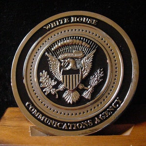 May include: A silver coin with a black background featuring the Presidential Seal of the United States and the text "White House Communications Agency".