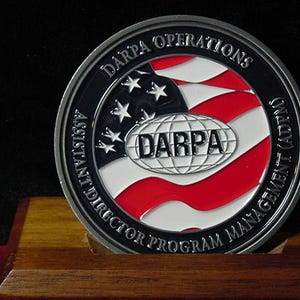 May include: A silver DARPA Operations coin with a red, white, and blue American flag design in the center. The coin features the DARPA logo and text around the edge, including "Assistant Director Program Management (ADPM)". The coin is displayed on a wooden base.