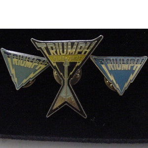 May include: Three metal pins featuring the word "Triumph" in a triangular shape. The center pin has a gold and yellow color scheme with a guitar pick design. The left and right pins have a blue and silver color scheme with a lightning bolt design.