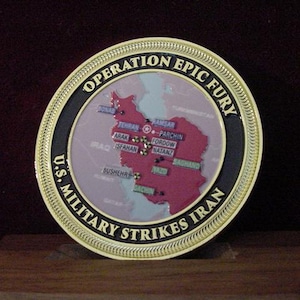 May include: A commemorative coin with a gold rim and black border. The coin features a map of Iran in red, with text that reads "OPERATION EPIC FURY" and "U.S. MILITARY STRIKES IRAN". The coin is displayed on a wooden surface.