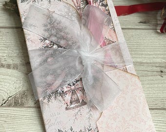 Può includere: Una busta di carta rettangolare con una stampa natalizia rosa pallido e bianca di stelle di Natale, alberi di pino e campane. Il testo recita All is Calm, All is Bright. Un nastro di organza bianco trasparente è legato a fiocco.