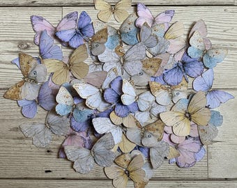 Può includere: Una collezione di farfalle di carta in morbide tonalità pastello, tra cui lavanda, azzurro e crema. Le farfalle presentano intricati disegni alari e sono disposte su una superficie di legno, creando un'esposizione artistica.