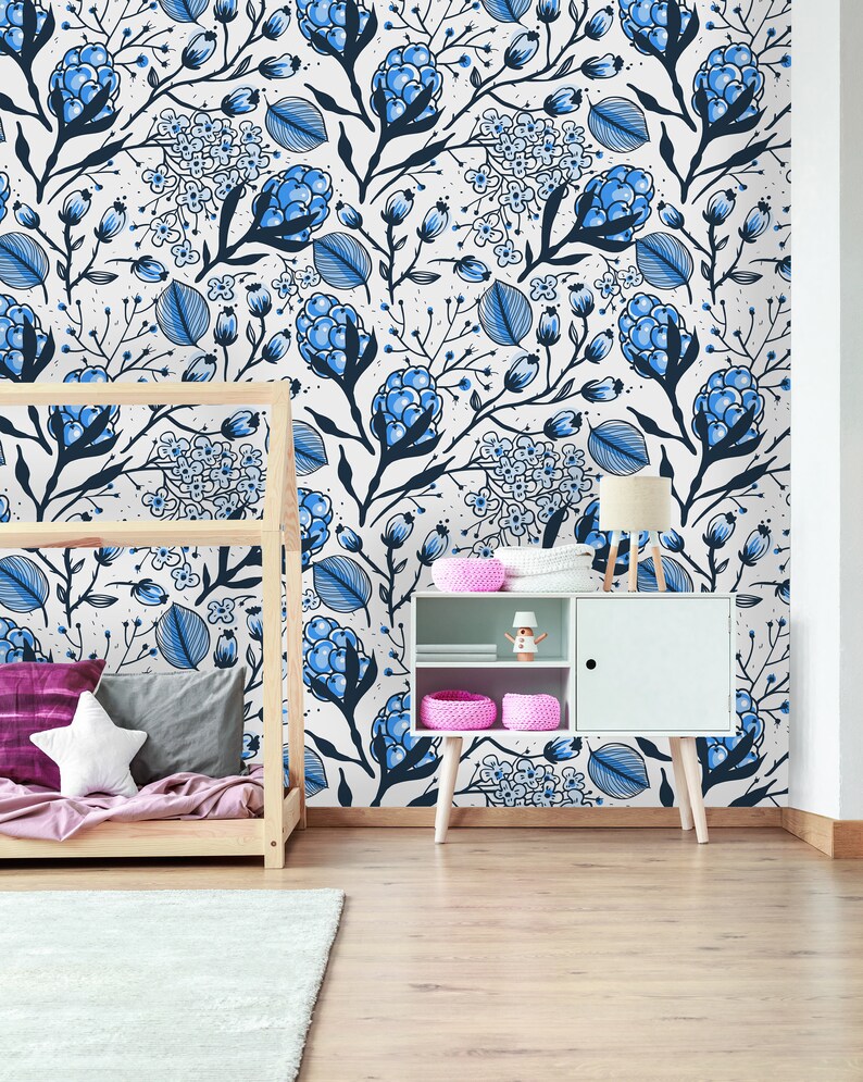 Puede incluir: Una pared blanca con un patr&oacute;n de papel tapiz floral azul y blanco. Un marco de cama de madera con una manta rosa y morada est&aacute; en primer plano. Un gabinete blanco con un estante y una cesta rosa est&aacute; en primer plano. Una l&aacute;mpara est&aacute; en el gabinete.