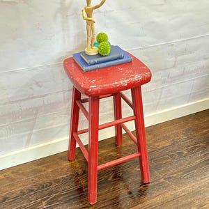 Pu&ograve; includere: Un piccolo sgabello in legno rosso con seduta quadrata e quattro gambe. Lo sgabello ha una finitura anticata. Sopra lo sgabello ci sono due libri blu, due palline decorative verdi e un manichino di artista in legno.