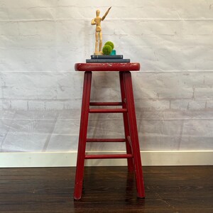 Pu&ograve; includere: Uno sgabello in legno rosso usurato con tre pioli. Lo sgabello &egrave; sormontato da libri, sfere di muschio decorative e un piccolo manichino di artista in legno. Lo sgabello &egrave; alto circa 61 cm.