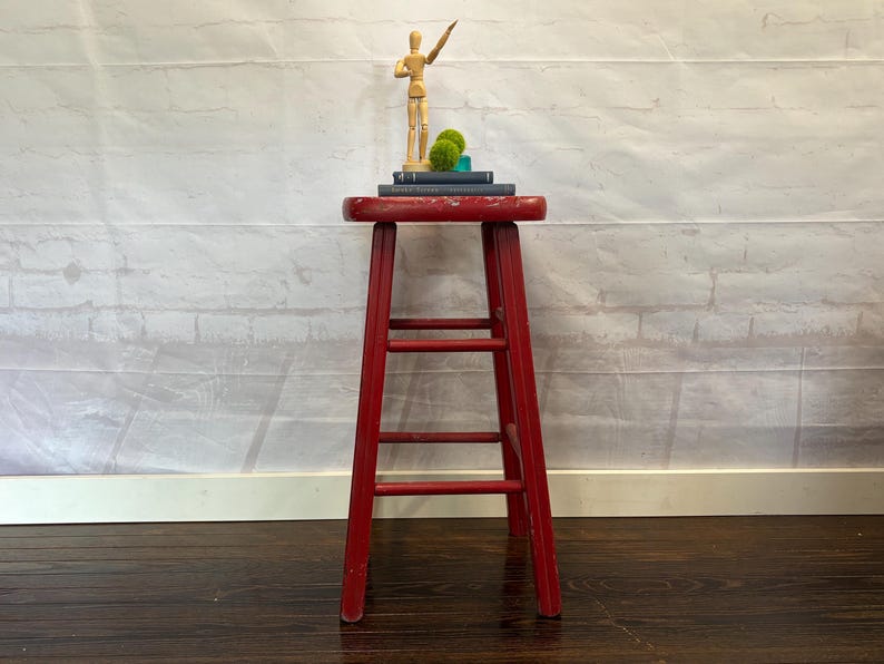 Pu&ograve; includere: Uno sgabello in legno rosso usurato con tre pioli. Lo sgabello &egrave; sormontato da libri, sfere di muschio decorative e un piccolo manichino di artista in legno. Lo sgabello &egrave; alto circa 61 cm.