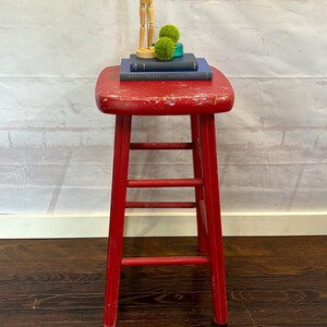 Pu&ograve; includere: Uno sgabello in legno rosso usurato con tre pioli e un piano quadrato. Sopra ci sono due libri blu scuro, due palline decorative verdi e un manichino di artista in legno. Lo sgabello &egrave; su un pavimento in legno scuro.