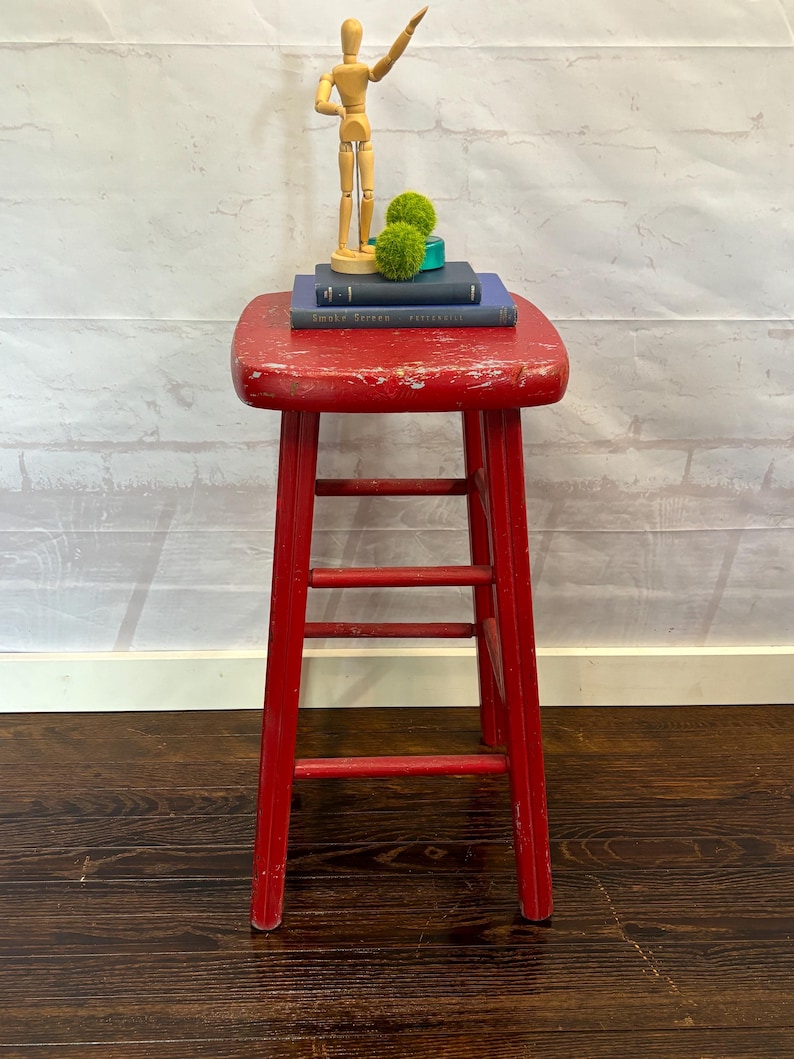 Pu&ograve; includere: Uno sgabello in legno rosso usurato con tre pioli e un piano quadrato. Sopra ci sono due libri blu scuro, due palline decorative verdi e un manichino di artista in legno. Lo sgabello &egrave; su un pavimento in legno scuro.