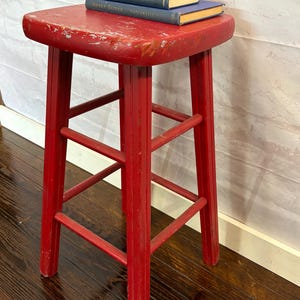 Pu&ograve; includere: Uno sgabello in legno rosso usurato con seduta quadrata e quattro gambe. Lo sgabello ha un aspetto vintage e usurato. Due piccoli libri e sfere verdi decorative sono appoggiati sopra.
