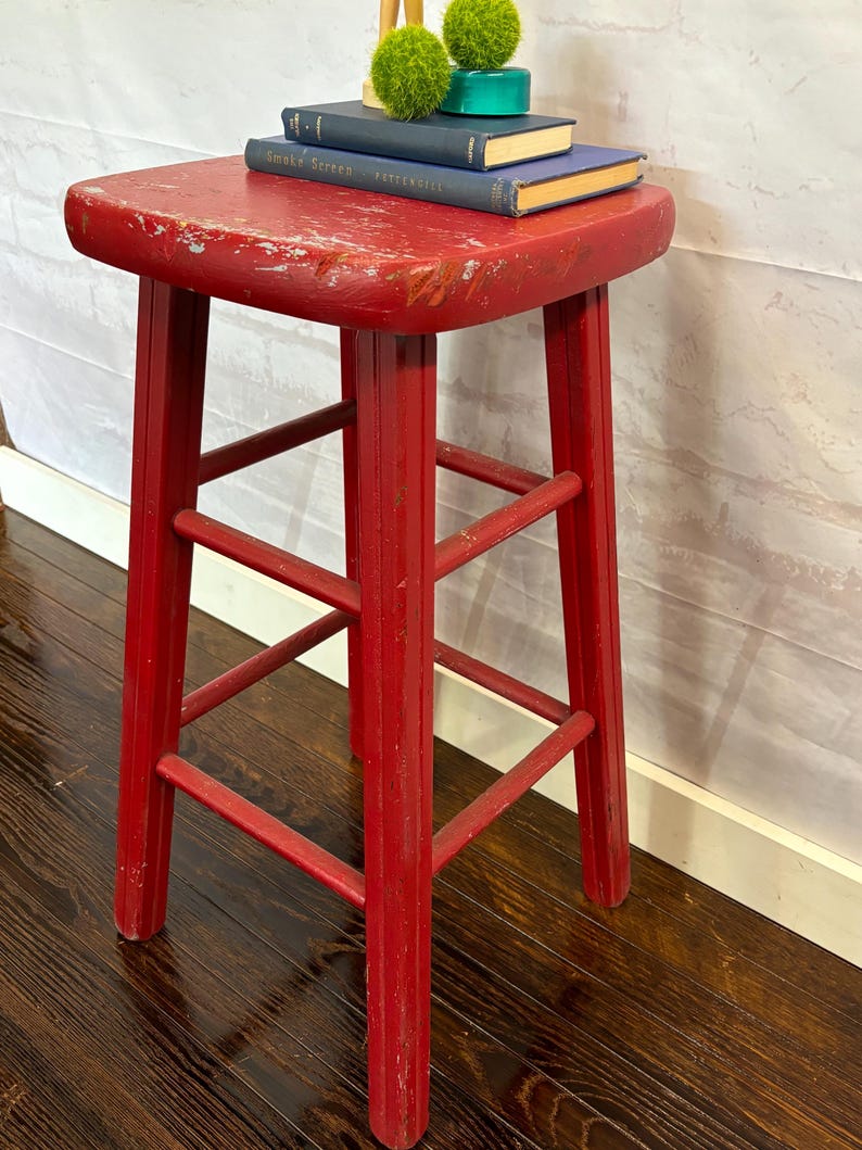 Pu&ograve; includere: Uno sgabello in legno rosso usurato con seduta quadrata e quattro gambe. Lo sgabello ha un aspetto vintage e usurato. Due piccoli libri e sfere verdi decorative sono appoggiati sopra.
