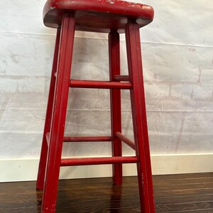 Pu&ograve; includere: Uno sgabello alto in legno rosso con seduta rettangolare e quattro gambe. Lo sgabello ha tre barre di supporto orizzontali. La vernice rossa &egrave; usurata, conferendogli un aspetto vintage. Un piccolo manichino di artista in legno e un pompon verde sono sopra.