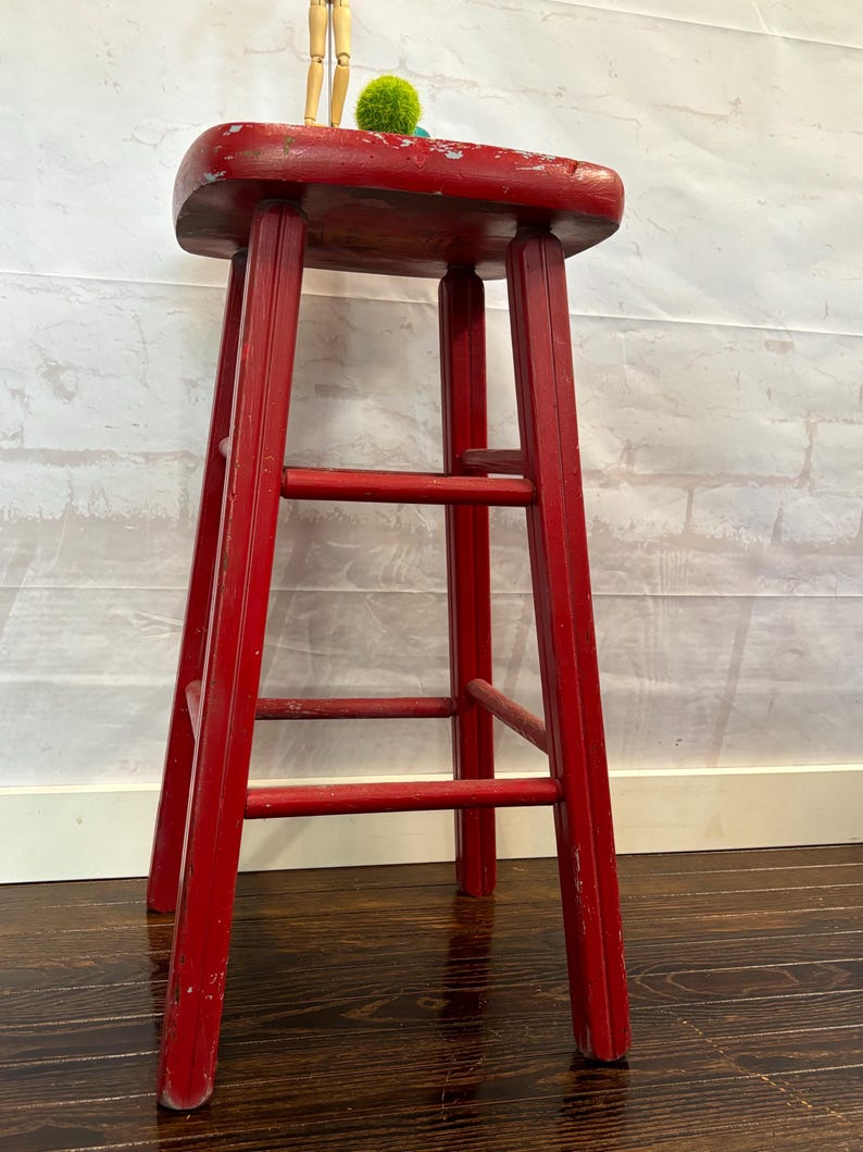 Pu&ograve; includere: Uno sgabello alto in legno rosso con seduta rettangolare e quattro gambe. Lo sgabello ha tre barre di supporto orizzontali. La vernice rossa &egrave; usurata, conferendogli un aspetto vintage. Un piccolo manichino di artista in legno e un pompon verde sono sopra.