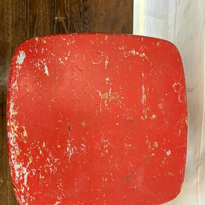 Pu&ograve; includere: Primo piano di un oggetto rosso di forma quadrata con angoli arrotondati. La vernice rossa &egrave; usurata, mostrando macchie bianche e dorate sottostanti. L'oggetto sembra essere di legno.