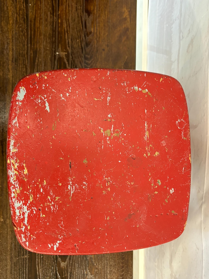 Pu&ograve; includere: Primo piano di un oggetto rosso di forma quadrata con angoli arrotondati. La vernice rossa &egrave; usurata, mostrando macchie bianche e dorate sottostanti. L'oggetto sembra essere di legno.