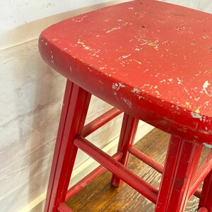 Pu&ograve; includere: Sgabello vintage in legno rosso con seduta quadrata e quattro gambe. Lo sgabello ha una finitura usurata, con segni di usura visibili sulla superficie verniciata. Le gambe sono collegate da supporti orizzontali.
