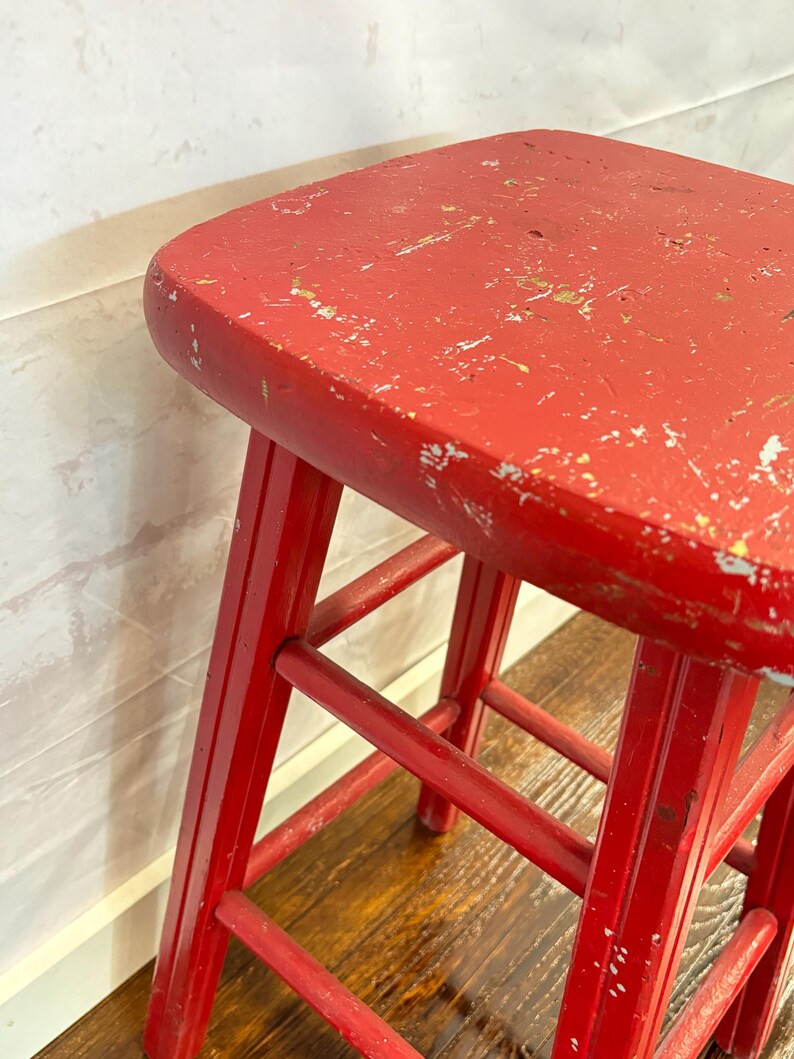 Pu&ograve; includere: Sgabello vintage in legno rosso con seduta quadrata e quattro gambe. Lo sgabello ha una finitura usurata, con segni di usura visibili sulla superficie verniciata. Le gambe sono collegate da supporti orizzontali.