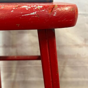 Pu&ograve; includere: Primo piano di uno sgabello in legno rosso, con segni di usura. Lo sgabello ha un sedile rotondo e quattro gambe. La vernice rossa &egrave; scheggiata, rivelando il legno sottostante. Due libri blu sono impilati sullo sgabello.