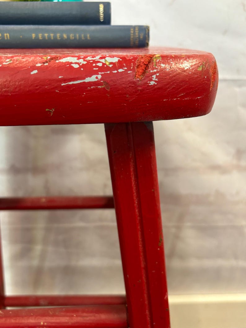 Pu&ograve; includere: Primo piano di uno sgabello in legno rosso, con segni di usura. Lo sgabello ha un sedile rotondo e quattro gambe. La vernice rossa &egrave; scheggiata, rivelando il legno sottostante. Due libri blu sono impilati sullo sgabello.