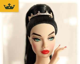 May include: A porcelain doll with long black hair, wearing a gold tiara with rhinestones. She has bright blue eyes and red lipstick. The doll is posing with her hand on her cheek.