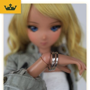 Puede incluir: Una muñeca con cabello rubio y ojos azules lleva una pulsera plateada en la muñeca.