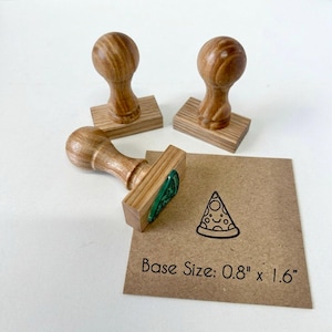 Könnte beinhalten: Drei Holzstempel mit abgerundeten Griffen und rechteckigen Sockeln. Ein Stempel liegt auf einer braunen Karte mit einem schwarzen Stempelabdruck eines lächelnden Pizzastücks und dem Text Base Size: 2,03 cm x 4,06 cm.
