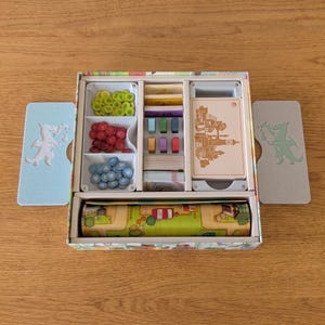 May include: A board game box organizer containing various game components including green, red, and blue circular tokens, small rectangular colored blocks, card decks, and a rolled game board. Two light blue and grey dragon-embossed side panels.