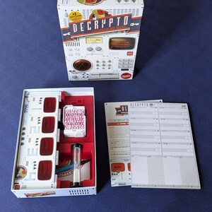 May include: A board game box and components are displayed on a blue surface. The box is white with red and black accents and the text "DECRYPTO". Inside the box are game pieces, including cards, a timer, and a game board.