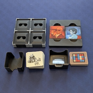 Op de afbeelding: Een verzameling bordspelcomponenten, waaronder zwarte plastic houders, speelkaarten en tegels. De tegels bevatten afbeeldingen van personages en een puzzel. De kaarten hebben illustraties van dieren en figuren.