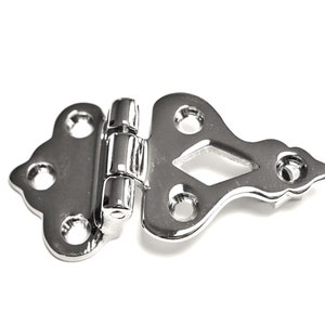 May include: A silver-toned decorative hinge with a diamond-shaped cutout in the center. The hinge has a decorative, ornate design and is made of metal.