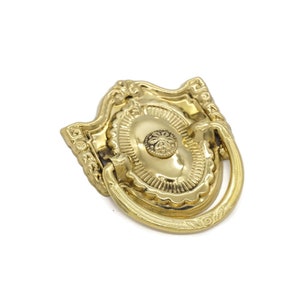 May include: Gold-toned ornate cabinet pull with a ring handle. The pull has a decorative, detailed design.