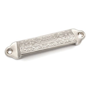 May include: A silver-toned, rectangular drawer pull with an ornate, engraved design. The pull has rounded edges and two small holes for mounting.