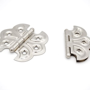 May include: Two silver butterfly-shaped hinges with a decorative design. The hinges are closed and have a polished finish.