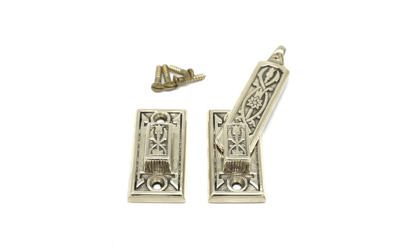 May include: Two antique brass door latches with ornate floral and geometric designs. The set includes two rectangular base plates, a pivoting latch, and several small brass screws. The hardware is set against a white background.