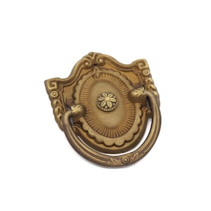 May include: A gold-toned metal cabinet pull with a decorative, ornate design. The pull features a ring handle and a central floral motif.