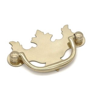 May include: A gold-toned metal cabinet pull with a leaf design. The pull has a curved handle and two round knobs.